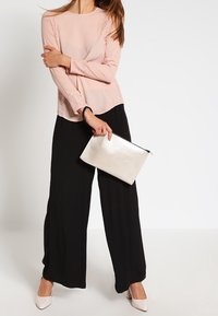 Femme portant un haut à manches longues rose avec des détails en dentelle, un pantalon noir à jambes larges, des talons blancs, tenant une pochette argentée.