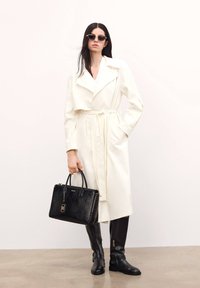 Fehér trench kabát derékra kötött övvel, fekete krokodilbőr textúrájú táska, és térd feletties fekete csizmák cipzárral. A modellen sötét napszemüveg van.