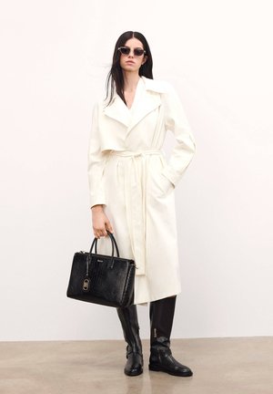 White trench coat with a tie waist, black crocodile-textured handbag, and knee-high black boots with zippers. Model wears dark sunglasses.