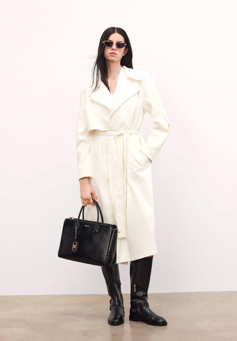Fehér trench kabát derékra kötött övvel, fekete krokodilbőr textúrájú táska, és térd feletties fekete csizmák cipzárral. A modellen sötét napszemüveg van.