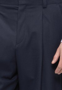 Κοντινό πλάνο σκούρων ναυτικών παντελονιών με μέση, ορατή μπροστινή ραφή και το δεξί χέρι ενός ατόμου που ακουμπά δίπλα στο ισχίο.