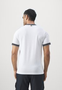 Polo-shirt blanc à manches courtes en tissu maillé texturé. Présente des rayures noires sur le col et les manches, ainsi qu'une étiquette colorée au niveau du cou.