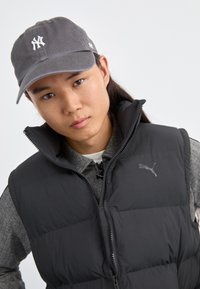 Gilet matelassé noir avec fermeture éclair à l'avant et col haut, porté par-dessus un pull gris à motifs, associé à une casquette de baseball grise arborant un logo blanc.
