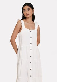 Abito bianco senza maniche con scollo quadrato, spalline arricciate, design a strati e bottoni marroni davanti, realizzato in un tessuto leggero.