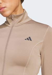 Beige Sportjacke mit Reißverschluss aus glattem Stoff, hohem Kragen, schwarzem Logo und dezenten Nahtdetails.