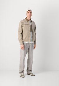 Chaqueta beige clara con un cuello marrón, capas sobre una camisa gris y unos jeans grises. Zapatillas marrones y verdes completan el atuendo.