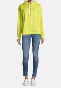 En person i en ljusgul huvtröja, smala blå jeans och vita sneakers, stående mot en neutral ljus bakgrund.