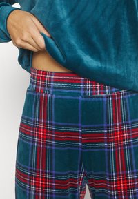 Main levant un haut en velours teal pour montrer un pantalon taille haute à motif écossais rouge, bleu, vert et blanc.