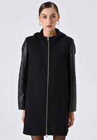Cappotto nero con zip frontale, maniche in pelle trapuntata e cappuccio. Texture liscia con un design moderno e minimalista.