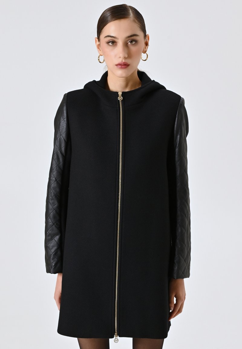 Cappotto nero con zip frontale, maniche in pelle trapuntata e cappuccio. Texture liscia con un design moderno e minimalista.