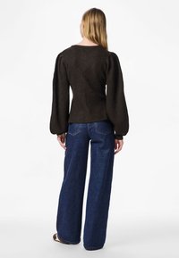 Femeie cu păr blond lung, văzută de spate, purtând un pulover maro închis cu mâneci bufante și blugi evazați albaștri, pe un fundal alb.