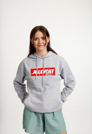 Junge Frau mit braunen Haaren, die einen grauen Kapuzenpullover mit rotem und weißem Schriftzug "ALLEY CAT" und grün gestreifte Shorts trägt, steht vor weißem Hintergrund.