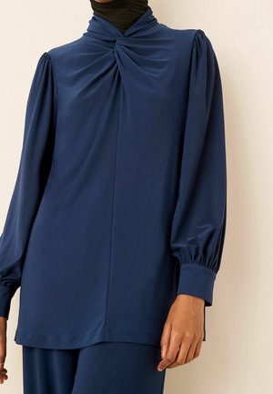 Personne portant un haut bleu marine à manches longues avec tissu froncé au niveau de l'encolure et manches bouffantes avec larges poignets.