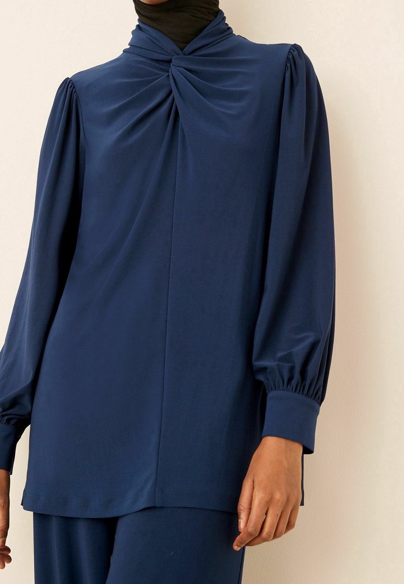 Personne portant un haut bleu marine à manches longues avec tissu froncé au niveau de l'encolure et manches bouffantes avec larges poignets.