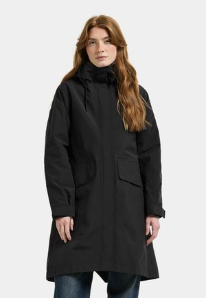 Vrouw met lang rood haar die een zwarte kapmantel met voorzakken en een blauwe spijkerbroek draagt, staand tegen een effen achtergrond.