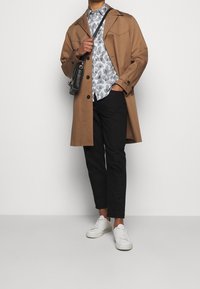 Ruskea trenssitakki kuvioidun harmaan paidan, mustien housujen ja valkoisten lenkkarien päällä. Mukana pieni musta olkalaukku.