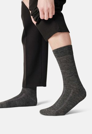 Graue, strukturierte Socken mit einem gerippten Bund, getragen mit schwarzen Hosen. Sie verfügen über ein dezentes Muster auf dem Stoff und eine taillierte Form.