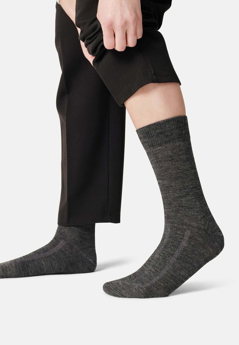 Graue, strukturierte Socken mit einem gerippten Bund, getragen mit schwarzen Hosen. Sie verfügen über ein dezentes Muster auf dem Stoff und eine taillierte Form.