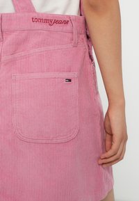 Rosa Corduroy-Latzhose mit verstellbaren Trägern, die eine hintere Tasche und ein kleines Markenetikett in Marineblau, Weiß und Rot am Taschenrand aufweist.