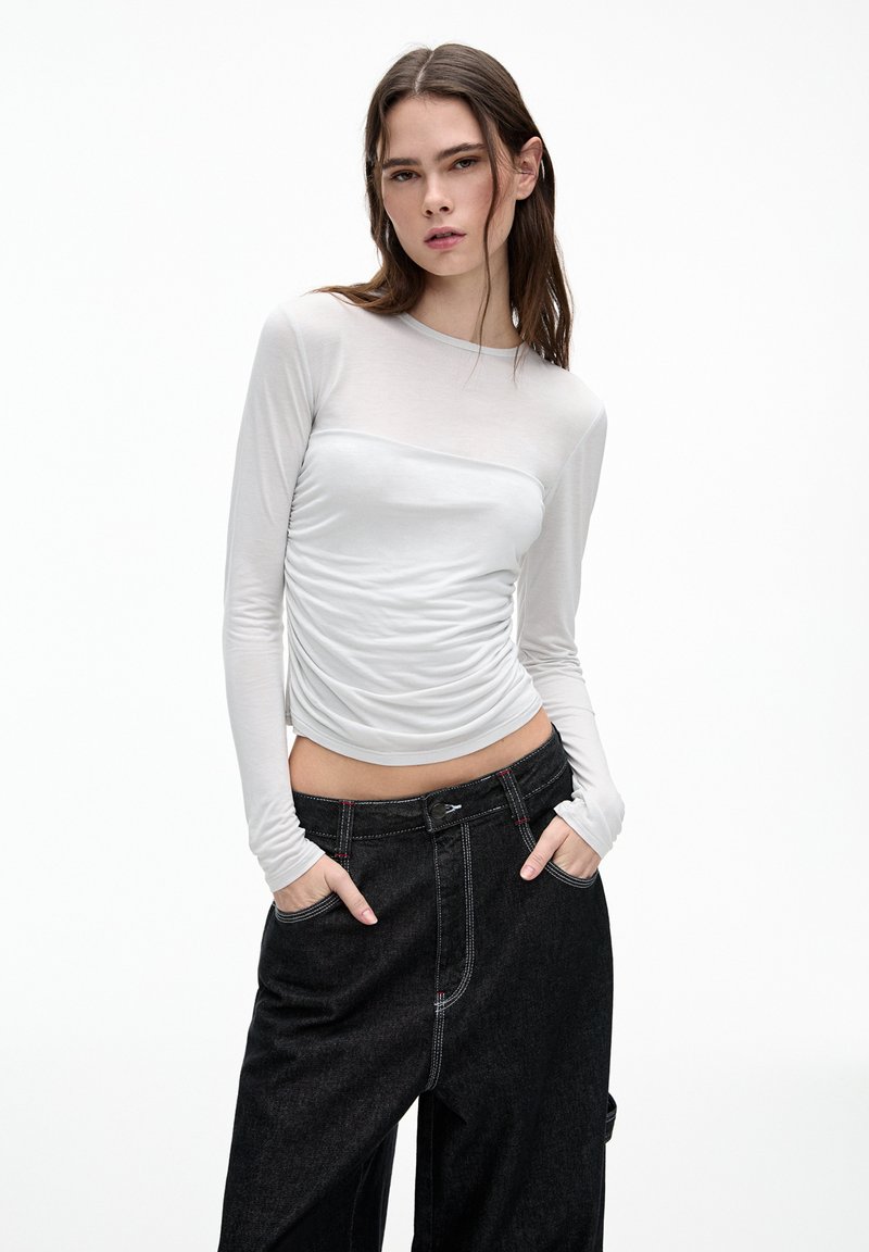Maglietta bianca a maniche lunghe con dettagli arricciati, realizzata in un tessuto leggero. Abbinata a jeans neri larghi con accenti di cucitura rossa.