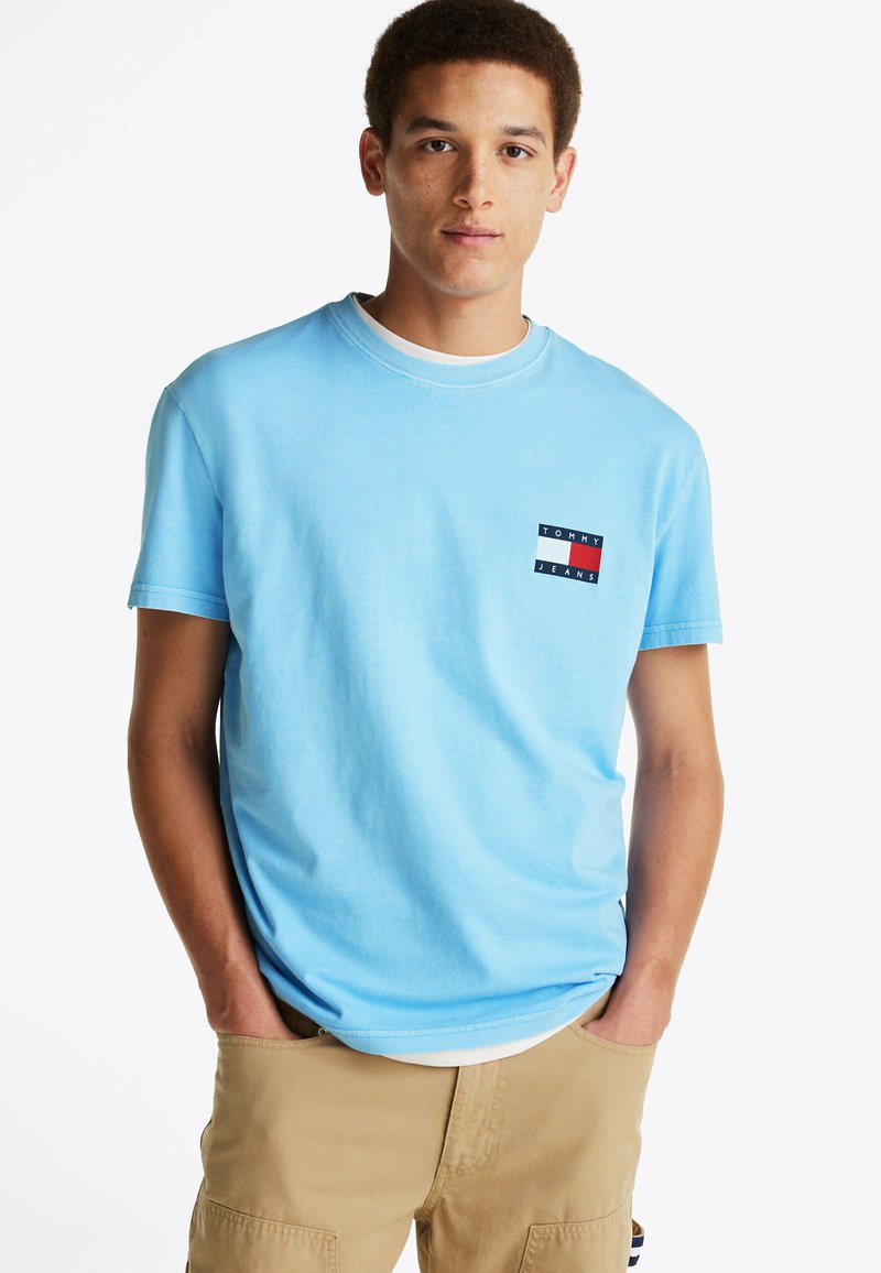 Camiseta de algodón azul claro con cuello redondo y mangas cortas. Presenta un pequeño parche con el logo en rojo, blanco y marino en el lado izquierdo del pecho.