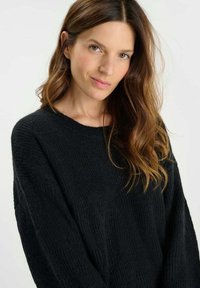 Maglione nero a maglia con vestibilità ampia e scollo largo. Presenta un tessuto a trama e bordi a coste. Il modello ha capelli mossi e un viso delicatamente incorniciato.