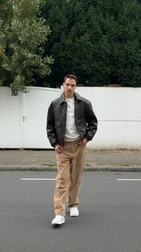 Veste en cuir marron, pull blanc, pantalon en velours côtelé beige et baskets blanches. Debout sur une rue avec un mur blanc et des arbres en arrière-plan.