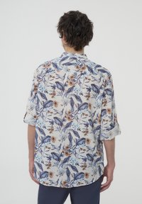 Camicetta a maniche corte con uno sfondo chiaro e motivi floreali blu, marrone e giallo. Il tessuto appare leggero con una vestibilità comoda.