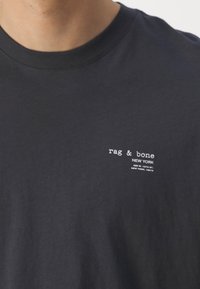 Musta puuvillainen t-paita, jossa on pyöreä kaula-aukko, ja vasemmassa rintakohdassa valkoinen etiketti, jossa lukee "rag & bone NEW YORK".