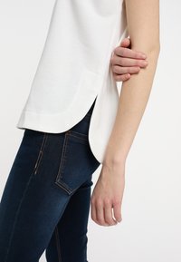 Bílý top bez rukávů s zakřiveným lemem a bočními rozparky, spárovaný s tmavě modrými džíny z denimu se zlatým prošíváním a přiléhavým střihem.