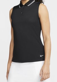 Černý bezrukávový polo top s bílým límečkem a třemi knoflíky, nošený s bílou sukni, která má blízko lemování malé bílé logo Nike.