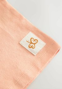 Tela de color durazno con una textura suave, que presenta una etiqueta blanca con un diseño de mariposa naranja en la esquina. Costuras visibles.