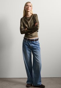 Olivgrünes langärmliges Oberteil, kombiniert mit blauen weiten Jeans und einem braunen Gürtel. Glatte Textur und lässiger Stil.