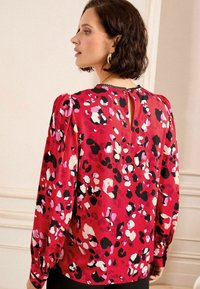 Blusa rossa con un motivo leopardato in nero, rosa e bianco. Presenta maniche lunghe, un'apertura a forma di chiave sul retro e spalle arricciate.