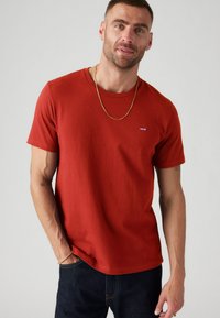 Tricou roșu din bumbac, cu mâneci scurte și decolteu rotund, având un mic logo al Levi's de culoare albastră pe piept și o croială normală. Este purtat cu blugi negri.