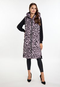 Langer, ärmelloser pinker Vest mit schwarzem Leopardenmuster, ausgestattet mit einem hohen Kragen und einem Reißverschluss vorne. Kombiniert mit schwarzen Leggings und spitzen Absätzen.