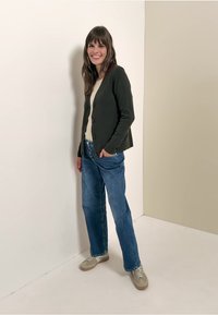 Mörkgrön cardigan över en ljus tröja, kombinerad med avslappnade blå jeans och neutrala sneakers. Enkelt mönster med knappdetaljer.