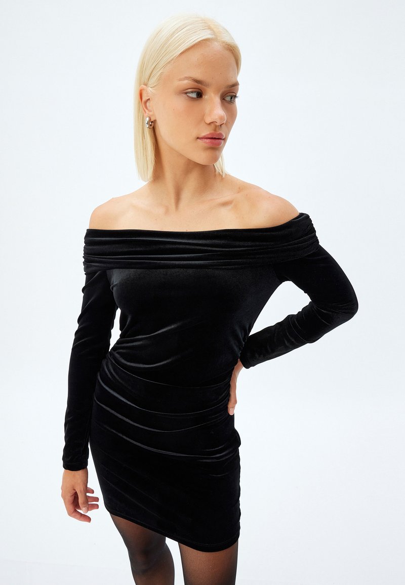 Zwart fluwelen off-shoulder jurk met lange mouwen, aansluitende silhoe t en geplooide details langs de taille en rok.