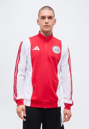 Homme portant une veste de sport Adidas rouge et blanche avec le logo de l'équipe sur la poitrine, debout sur un fond clair uni.