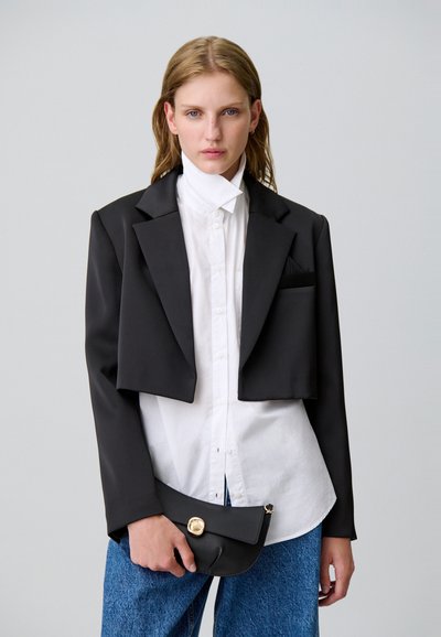 Blazer negro recortado con textura suave y cuello chal, combinado con una camisa blanca abotonada y un bolso de mano negro con un detalle dorado.