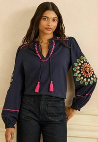 Navyblauwe blouse met geborduurde ontwerpen in levendige kleuren, voorzien van pofmouwen, een V-hals en roze kwastjes aan de koorden.