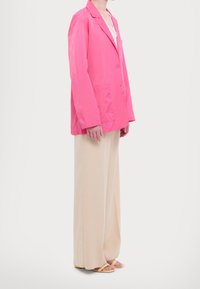 Blazer léger rose vif avec grandes poches, associé à un pantalon beige à jambes larges. Le modèle porte des sandales minimalistes avec des brides roses et jaunes.