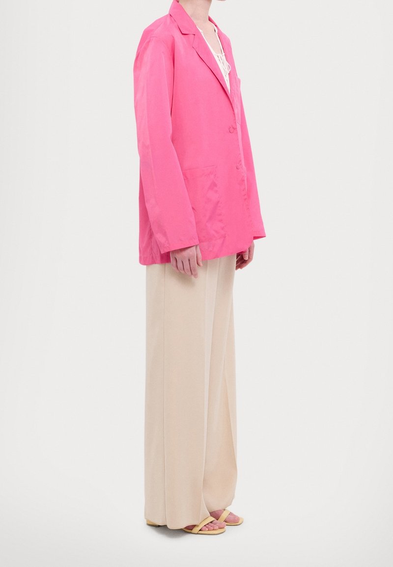 Blazer léger rose vif avec grandes poches, associé à un pantalon beige à jambes larges. Le modèle porte des sandales minimalistes avec des brides roses et jaunes.