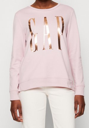 Personne portant un sweat-shirt à manches longues rose clair avec des lettres « GAP » en or rose brillant et un pantalon blanc, debout devant un fond blanc uni.