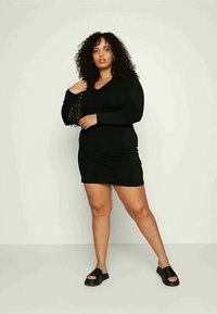 Vestido preto de manga longa com decote em V e detalhes de franzido lateral. Modelo usando sandálias pretas com salto plano, com cabelo cacheado solto.