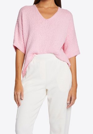 Kvinde iført en løs, kortærmet pink striktop, der er stoppet ned i højtaljede hvide bukser mod en hvid baggrund.