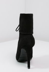 Černé semišové vysoké podpatky kotníkové boty s ohnutým lemem a stiletto podpatkem, zobrazené ze zadní strany na bílé ploše.