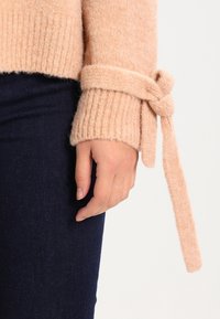 Mjuk rosa stickad jumper med ribbade ärmslut, med ett knytband vid handleden. Bärs över mörka denimjeans.