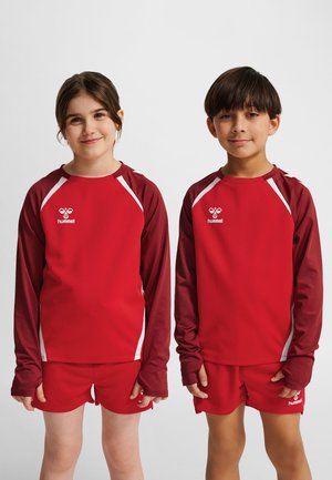 Deux enfants debout côte à côte, portant une tenue de sport rouge assortie avec des manches longues et un short, sur un fond uni.