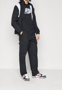 Černá nylonová bunda s bílými akcenty, černé tričko s potiskem, černé cargo kalhoty a černo-červené tenisky s bílou podrážkou.